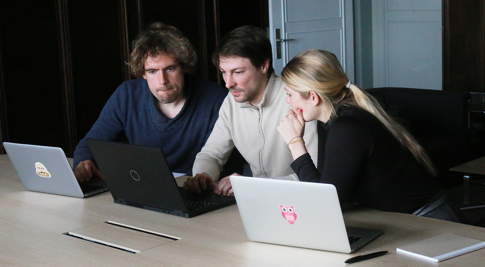 Zwei Männer und eine Frau sitzen an einem Tisch mit Laptops in einer Arbeitssituation